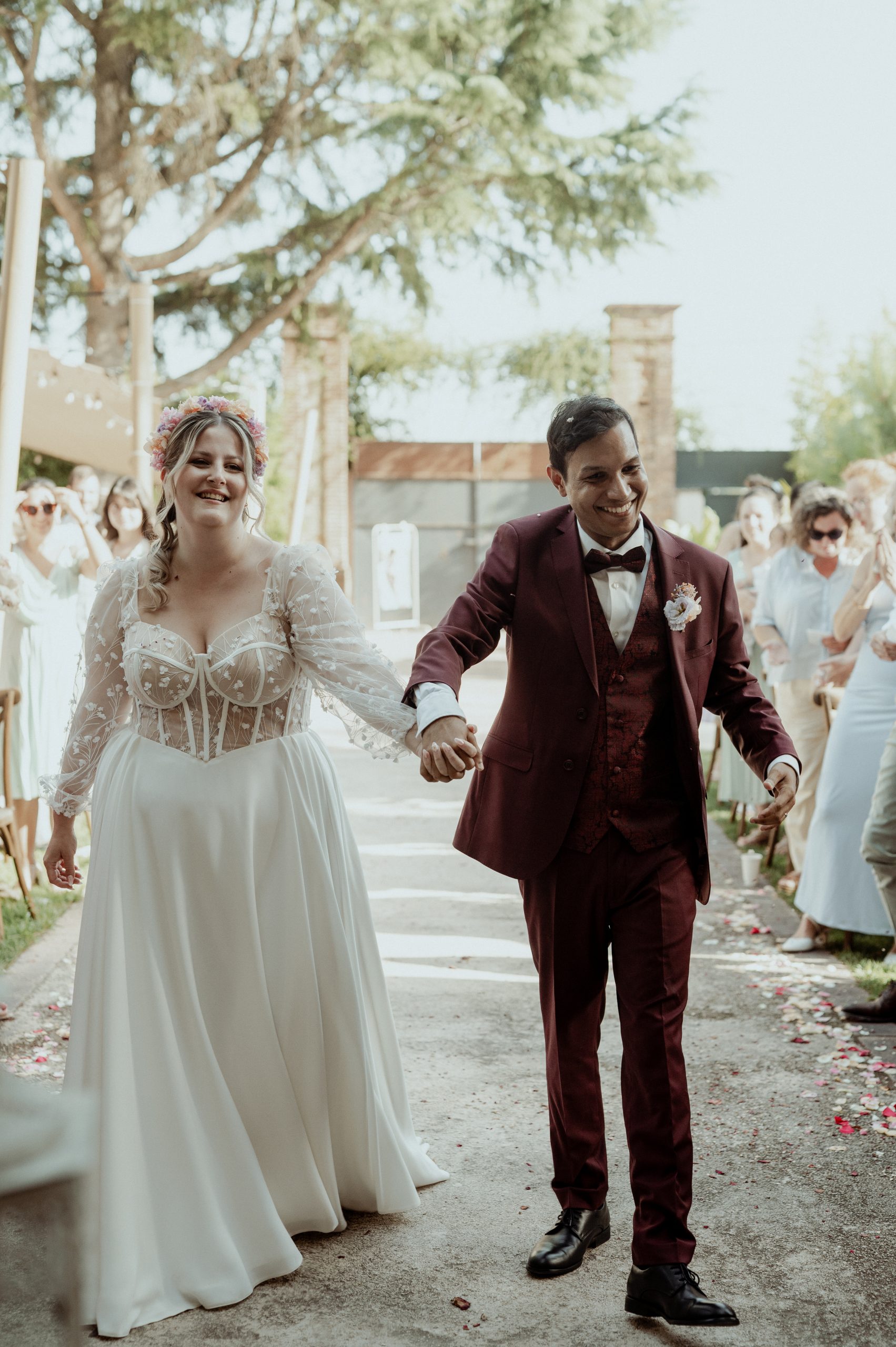 Emma et Maël avancent main dans la main lors de leur mariage. Emma porte une robe de mariée sur mesure avec un corset brodé et une jupe fluide.
