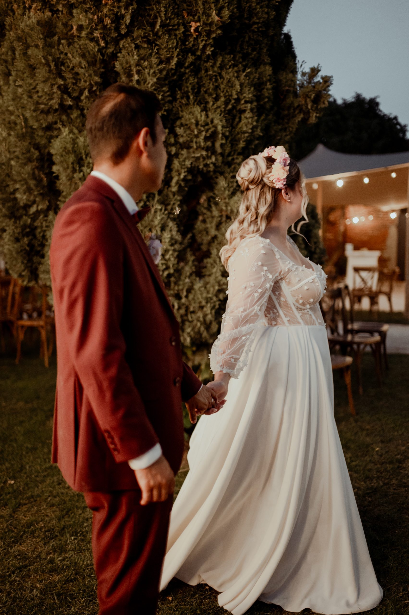 Emma et Maël se tiennent la main lors de leur mariage. Sa robe de mariée sur mesure associe un corset en tulle brodé à une jupe fluide avec des plis.